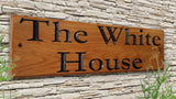 An LD Trade Personalised Oak House Number Name Sign, carved and engraved from European Oak, elegantly displays the words "The White House" in Garamond font, mounted on a stone wall. A layer of polyurethane varnish provides a subtle sheen while green plants thrive in the foreground.