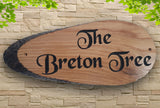 A personalized oak carved wooden house sign plaque, crafted by LD Trade with live edges, is mounted on a stone wall and displays "The Breton Tree" in elegant black text. Framed by green leaves at the top corners, its enduring beauty is preserved by a high-quality polyurethane varnish.