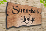 The LD Trade Personalised Oak Carved Wooden Sign House Plaque Outdoor, made from European Oak and featuring decorative black lettering that reads "Sunnybank Lodge," is coated in a protective polyurethane varnish. It is mounted on a light stone wall with greenery adorning the top corners.