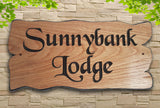 A personalised carved wooden sign house plaque from LD Trade, crafted from European Oak, is mounted against a stone wall and reads "Sunnybank Lodge" in bold, black script. The sign is finished with a protective polyurethane varnish and is artistically framed by green leaves at the top corners of the image.