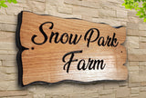 A beautifully crafted wooden sign from LD Trade, made from European Oak, features the text "Snow Park Farm" in elegant black script. This personalised house plaque is mounted on a stone wall and protected by a glossy layer of polyurethane varnish, while green leaves peek from the top corners, reflecting its picturesque outdoor setting.