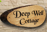 A "Personalised Oak Carved Wooden House Sign Plaque Slice Live edges" by LD Trade, featuring the words "Deep Well Cottage" elegantly engraved in Black Chancery Font, is mounted on a stone wall. Its natural, rustic charm is accentuated by a dark border and protected with a high-quality polyurethane varnish for lasting durability.