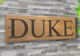 A Personalised Oak House Sign by LD Trade, featuring the word "DUKE" custom engraved in bold black letters, is mounted against a stone wall backdrop. Its polyurethane varnish finish has a subtle gleam, enhanced by green leaves adorning the upper right corner.