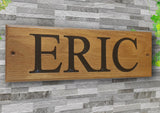 The Personalised Oak House Sign by LD Trade is a custom engraved, wooden name plaque crafted from European Oak, featuring the name "ERIC" in large, black letters. This elegant piece is mounted on a textured gray stone wall, with green vine leaves adding a natural touch in the corner.