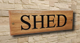 The LD Trade Personalised Oak House Sign, featuring the word "SHED" in bold black letters, is elegantly mounted on a light stone wall, showcasing a sleek polyurethane varnish finish.