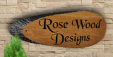 A beautifully carved wooden sign, made from high-quality European Oak, is prominently displayed on a textured stone wall. The sign elegantly features "Rose Wood Designs" in black script akin to Black Chancery Font. Its rich polyurethane varnish adds a lustrous finish, with a small green plant artistically positioned in the lower left corner of the scene.