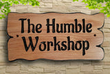 A bespoke Personalised Oak Carved Wooden Sign House Plaque Outdoor from LD Trade, featuring a protective layer of polyurethane varnish, displays the message "The Humble Workshop." It is stylishly installed on a stone wall with green leaves artistically framing the image at the top corners.