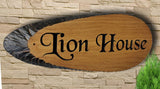 A personalized oak carved wooden house sign plaque slice with live edges, from LD Trade, featuring the text "Lion House" in elegant Black Chancery font, is mounted on a textured stone wall. A small section of greenery is visible in the bottom right corner, accentuating the high-quality polyurethane varnish that provides a polished finish.